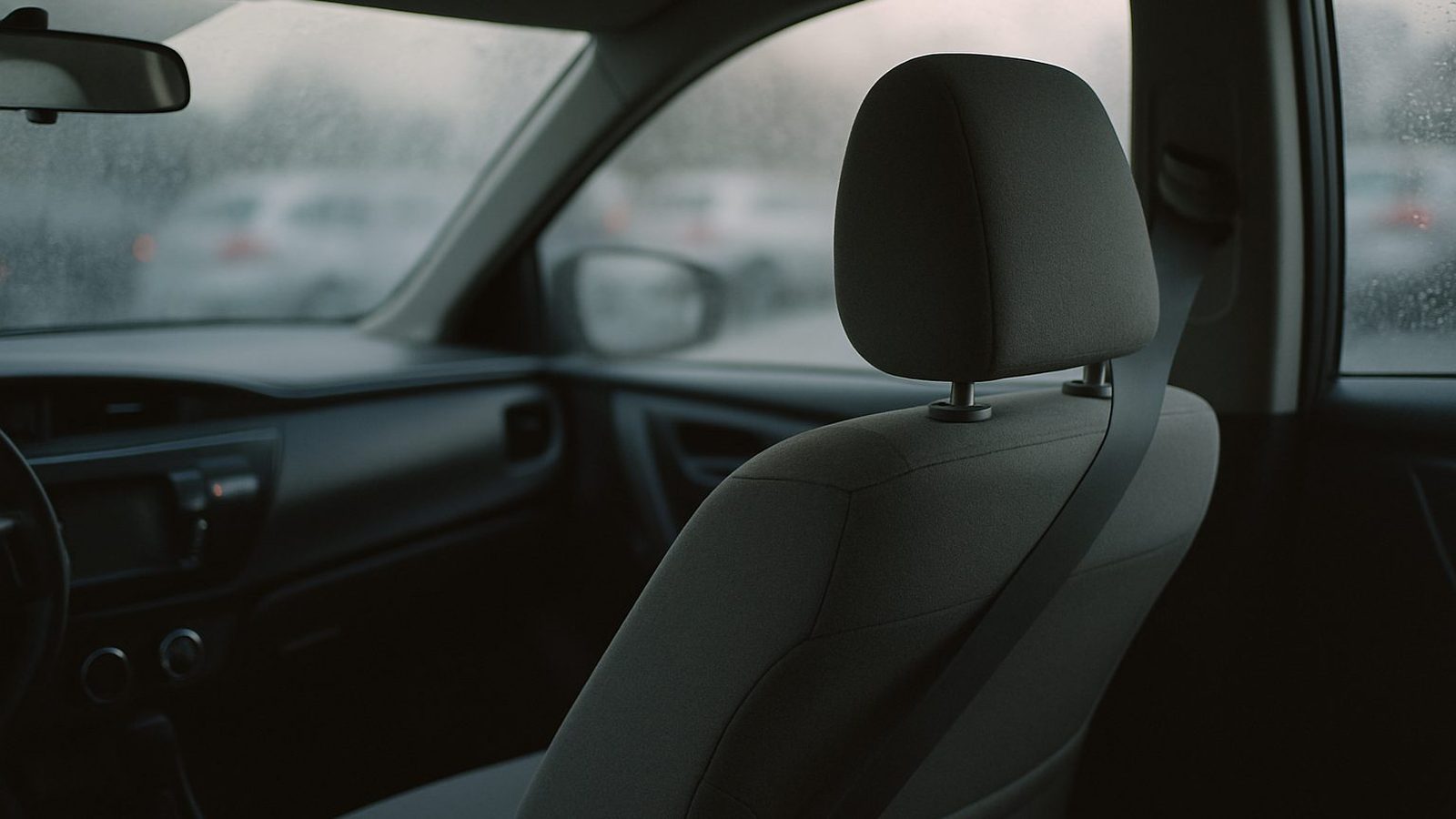 Inside a parked sedan on a grey morning, the driver's seat headrest and shoulder belt in focus, dew on the windshield, a parking lot blurred outside