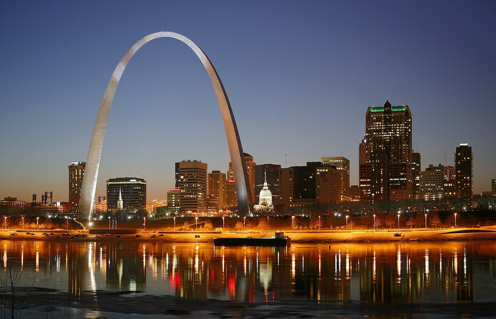 The Gateway Arch and the downtown St. Louis skyline at dusk, reflected in the Mississippi River with the riverfront lit in amber